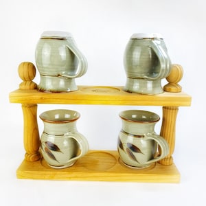 May include: A wooden mug rack with four ceramic mugs. The mugs are glazed in a light green colour with brown leaf designs. The rack has two shelves and wooden posts.