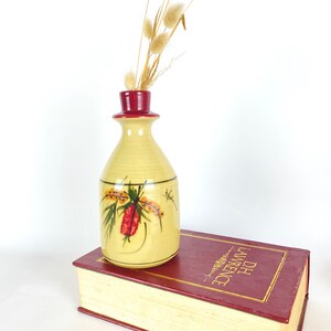 May include: A yellow ceramic vase with a red rim and a painted floral design. The vase is sitting on top of a red book with the title "D.H. Lawrence" on the spine.