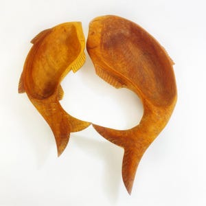 Op de afbeelding: Twee houten kommen in de vorm van een vis, elk met een gesneden ontwerp en een natuurlijke houten afwerking. De kommen zijn zo geplaatst dat ze naar elkaar toe wijzen, waardoor een decoratieve weergave ontstaat. Ideaal voor het serveren van snacks of als decoratieve stukken.