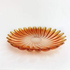 May include: An orange and gold glass decorative dish. The dish is oval-shaped with a flower petal design. The petals are orange with gold tips, and the centre is a lighter shade of orange.