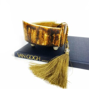 May include: A small, triangular ceramic bowl with a brown and white glaze. The bowl has three small feet and is sitting on a blue book with the title "VAN GOGH" printed on the cover. A gold tassel hangs from the bowl.
