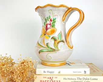 Op de afbeelding: Een witte keramische kan met een bloemenmotief in tinten geel, groen, rood en bruin. De kan heeft een gegolfde rand en een handvat. De kan staat op twee boeken, een met de titel "Be Happy" en de andere met de titel "The Art of Happiness".