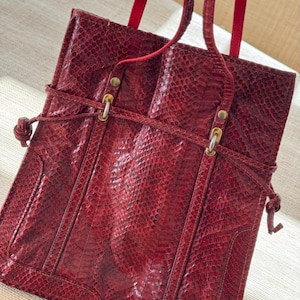 May include: A red snakeskin-textured tote bag with red straps. The bag has a rectangular shape with a flap closure and a braided leather detail. The bag has gold-colored hardware and a red interior.