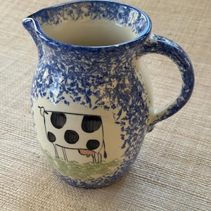May include: A ceramic pitcher with a blue speckled rim and handle. The body of the pitcher features a hand-painted cow illustration on a white background, with a green base. The pitcher has a spout for easy pouring.