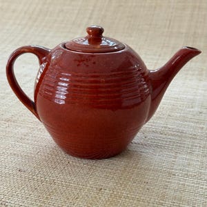 May include: A glossy, deep reddish-brown ceramic teapot featuring a rounded body with horizontal ribbed detailing. The teapot includes a matching lid with a small knob handle, a curved spout, and a side handle, all finished in a uniform glaze.