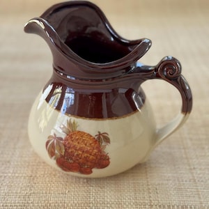 May include: A vintage ceramic pitcher with a brown rim and handle, and a cream-colored body. The pitcher features a pineapple and berry design. The handle has a decorative swirl. The pitcher is likely used for serving beverages.