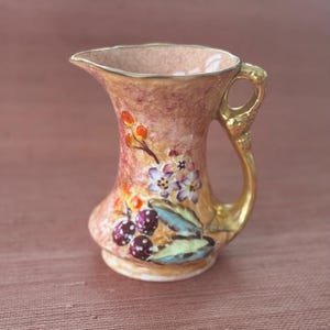 May include: A small, decorative pitcher with a flared top and a curved handle. The pitcher is painted with a floral design featuring berries and flowers in shades of red, purple, and blue. The rim and handle are gold-colored.