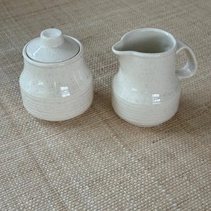 May include: A cream-colored ceramic sugar bowl with a matching lid and a coordinating creamer pitcher. Both pieces feature a speckled finish, a rounded body shape, and horizontal ribbed detailing around the base of each vessel.