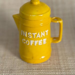 May include: A small, yellow coffee pot with a lid and handle. The words "INSTANT COFFEE" are printed in white on the front of the pot. The pot is made of metal and has a glossy finish.
