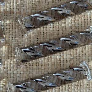 May include: Several clear glass, twisted-design drawer pulls are arranged on a woven, light brown surface. The pulls have a cylindrical shape with a spiral pattern and rounded ends. The glass reflects light, creating highlights and shadows.