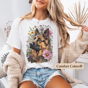 Puede incluir: Camiseta blanca con una ilustraci&oacute;n detallada de un perro pastor alem&aacute;n rodeado de flores coloridas. El perro tiene pelaje marr&oacute;n y negro. El texto "Comfort Colors&reg;" est&aacute; impreso en la parte inferior derecha.