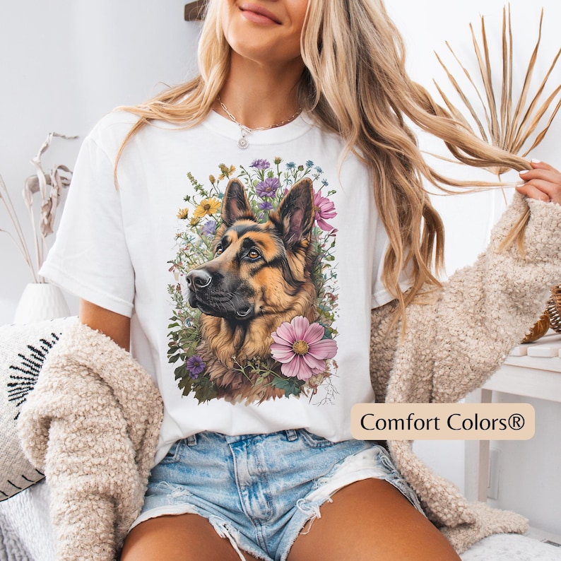 Puede incluir: Camiseta blanca con una ilustraci&oacute;n detallada de un perro pastor alem&aacute;n rodeado de flores coloridas. El perro tiene pelaje marr&oacute;n y negro. El texto "Comfort Colors&reg;" est&aacute; impreso en la parte inferior derecha.