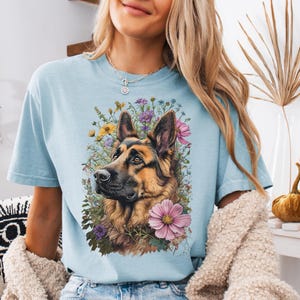 Puede incluir: Camiseta azul claro con un dise&ntilde;o de perro pastor alem&aacute;n rodeado de flores. El perro tiene pelaje marr&oacute;n y negro, y un aro en la nariz. Las flores son rosas, amarillas y moradas. La camiseta es de estilo casual y relajado.