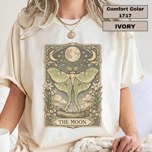 Puede incluir: Camiseta de color marfil con una ilustración detallada de una polilla, una luna llena y elementos celestiales. El gráfico está enmarcado con bordes ornamentados y el texto "THE MOON" en la parte inferior. La camiseta también tiene el texto "Comfort Color 1717 IVORY".