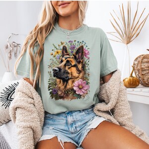 Puede incluir: Camiseta verde claro con una ilustraci&oacute;n detallada de un perro pastor alem&aacute;n rodeado de flores coloridas. El perro tiene pelaje marr&oacute;n y negro, y las flores incluyen flores rosas, moradas y amarillas. La camiseta es de corte holgado.