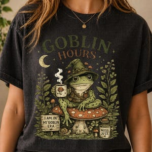 Puede incluir: Camiseta gris oscuro con una ilustración caprichosa. Una rana verde con sombrero y taza está sentada en una seta. La camiseta presenta el texto "GOBLIN HOURS" y "I AM IN MY GOBLIN ERA".