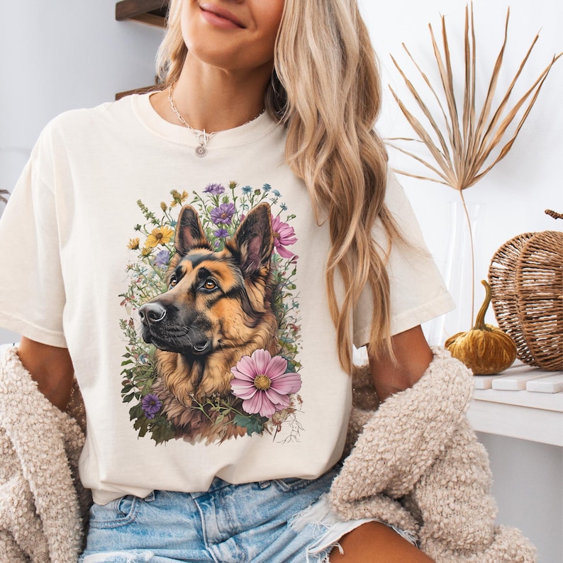 Puede incluir: Camiseta color crema con una ilustraci&oacute;n detallada de un perro pastor alem&aacute;n rodeado de flores coloridas. El pelaje del perro es marr&oacute;n y negro, con una mirada enfocada. Las flores incluyen flores rosas, amarillas y moradas, creando un dise&ntilde;o vibrante.