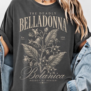 Puede incluir: Una camiseta gris carbón con una ilustración botánica de una planta de belladona con bayas y hojas. El texto THE DEADLY BELLADONNA, Est. 1753, Botanica y DEADLY BY DESIGN aparece en una mezcla de fuentes serif y cursiva en el centro del pecho.