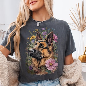 Puede incluir: Camiseta gris oscuro con un retrato de un perro pastor alem&aacute;n rodeado de flores coloridas. El perro tiene pelaje marr&oacute;n y negro, con un arreglo floral de flores rosas, moradas y amarillas. La camiseta tiene cuello redondo y mangas cortas.