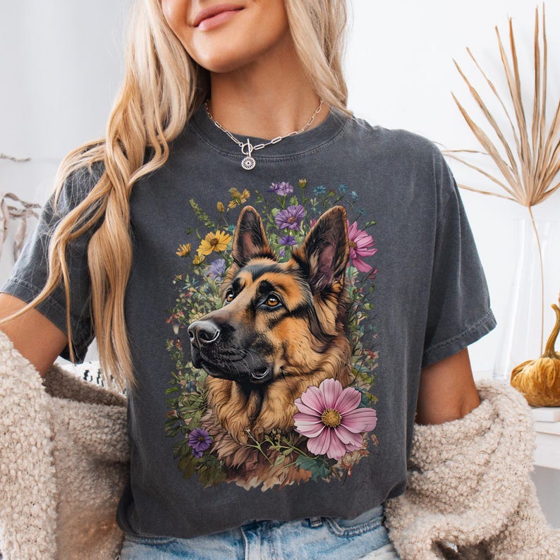Puede incluir: Camiseta gris oscuro con un retrato de un perro pastor alem&aacute;n rodeado de flores coloridas. El perro tiene pelaje marr&oacute;n y negro, con un arreglo floral de flores rosas, moradas y amarillas. La camiseta tiene cuello redondo y mangas cortas.