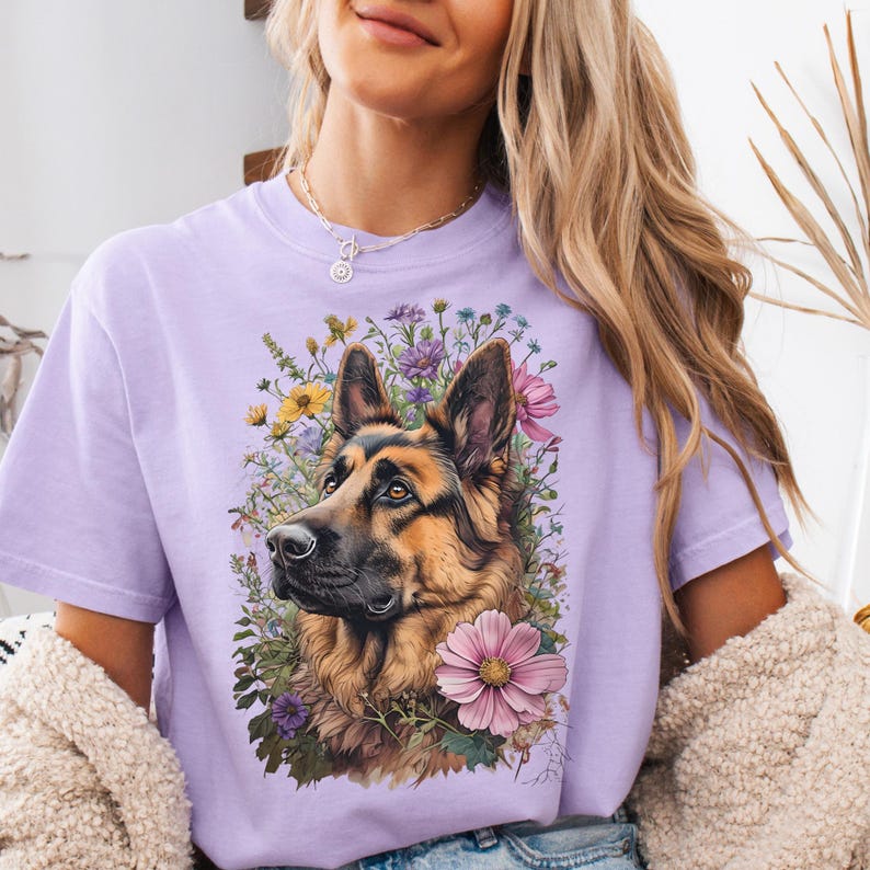 Puede incluir: Una camiseta de color lila con una ilustraci&oacute;n detallada de un perro pastor alem&aacute;n rodeado de flores coloridas. El perro tiene pelaje marr&oacute;n y negro, con una mirada enfocada. Las flores incluyen flores rosas, amarillas y moradas, creando un dise&ntilde;o vibrante.
