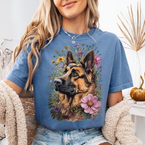 Puede incluir: Camiseta azul con un dise&ntilde;o gr&aacute;fico de un perro pastor alem&aacute;n rodeado de flores coloridas. El perro tiene pelaje marr&oacute;n y negro, con un arreglo floral detallado. La camiseta es de estilo casual.