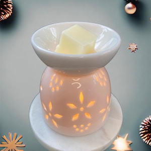 Op de afbeelding: Een witte keramische waxbrander met een lichtroze basis met bloemvormige uitsparingen. Een wit schaaltje bevat een lichtgele waxmelt. De brander staat op een witte marmeren basis, met decoratieve elementen.