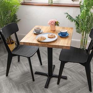 Puede incluir: Una mesa cuadrada de madera con una base de metal negro, dos sillas negras y un plato azul y blanco con dos pasteles. Hay dos tazas de café en la mesa, una en una taza azul y otra en una taza blanca. Un jarrón con flores rosas se encuentra en la mesa.