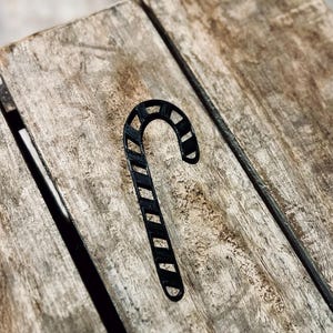 Könnte beinhalten: Eine schwarze, stilisierte Zuckerstangen-Dekoration mit Streifenmuster. Die Zuckerstange liegt auf einer verwitterten Holzoberfläche. Das Bild ist leicht von oben aufgenommen und hebt die Textur des Holzes und die Form der Zuckerstange hervor.