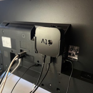May include: A black Apple TV streaming device is mounted on the back of a black television. The Apple TV is connected to the television with two black cables. There is a white cable connected to the television as well.