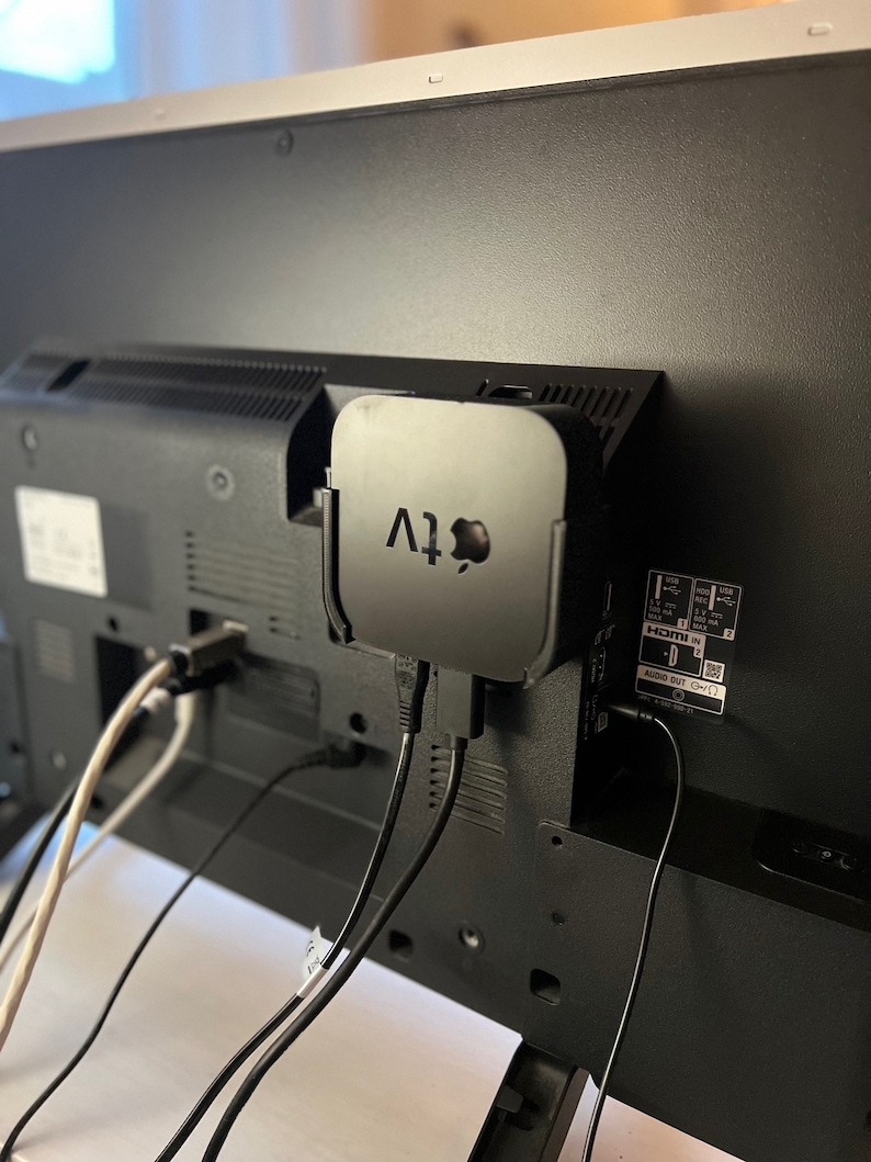 May include: A black Apple TV streaming device is mounted on the back of a black television. The Apple TV is connected to the television with two black cables. There is a white cable connected to the television as well.
