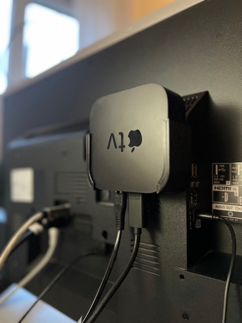 May include: A black Apple TV streaming device is mounted on the back of a black television. The device is connected to the television with two black cables. The television has a HDMI input and an audio output.