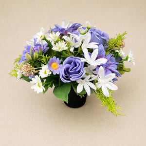Peut inclure: Un arrangement floral composé de fleurs artificielles aux tons de violet, blanc et vert. L'ensemble comprend des roses, des marguerites et d'autres fleurs, le tout disposé dans un petit pot noir. Les fleurs sont agencées pour un aspect plein et luxuriant.