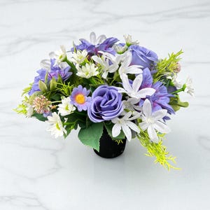 May include: A vibrant floral arrangement featuring purple roses, white star-shaped flowers, and small yellow-centered daisies. The artificial flowers are arranged in a black pot, creating a colorful and decorative display.