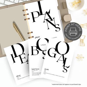 Könnte beinhalten: Ein Planer mit einem goldenen Stift und drei Karten mit den Wörtern "Ideen", "Lady Boss" und "Ziele". Der Planer ist auf einer Seite mit der Aufschrift "PLANS" geöffnet. Die Karten haben schwarzen Text auf weißem Hintergrund.