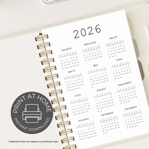 May include: A white 2026 calendar planner with gold spiral binding. The calendar pages display the months of the year in a simple, minimalist font. The planner includes the text "PRINT AT HOME" and "INSTANT DOWNLOAD".