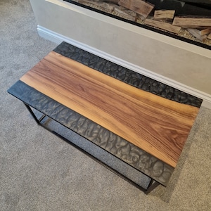 May include: A rectangular coffee table featuring a central panel of natural brown wood grain flanked by two textured black resin sections with a wavy, organic pattern. The table is supported by a thin, black metal frame with four legs.
