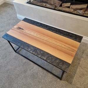 May include: A rectangular coffee table featuring a central light brown wood plank with a natural grain and dark knot detail, flanked by two side panels of dark grey swirling resin. The table is supported by a black metal frame base.