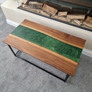May include: A rectangular coffee table featuring a natural brown wood grain surface divided by a central, swirling emerald green resin inlay. The tabletop is supported by a black metal frame with four straight legs and a rectangular base.