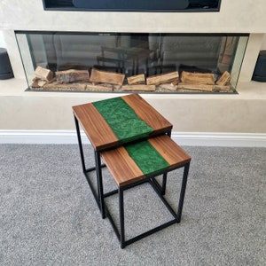 May include: A set of two nesting tables featuring rectangular walnut wood tops with a central river-like inlay of swirling emerald green resin. Each table is supported by a black metal frame with four straight legs and a square base design.