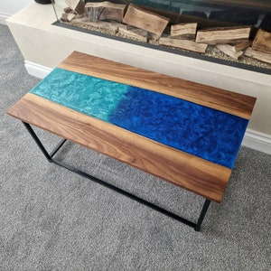 May include: A rectangular coffee table featuring two natural wood slabs with a central river inlay made of swirling resin. The resin transitions from a bright turquoise to a deep royal blue. The table is supported by a black metal frame base.