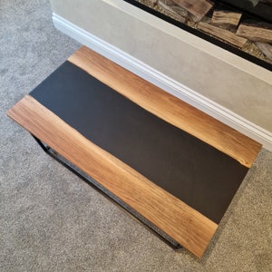 May include: A rectangular coffee table featuring two natural wood slabs with live edges flanking a wide, matte black resin centre inlay. The table is supported by a thin, black metal frame base, showcasing a contrast between the wood grain and resin.