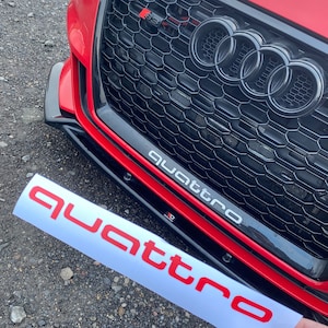 May include: A red Audi with a black grille and the word "quattro" written in white on the front bumper. A red "quattro" sticker is shown separately.
