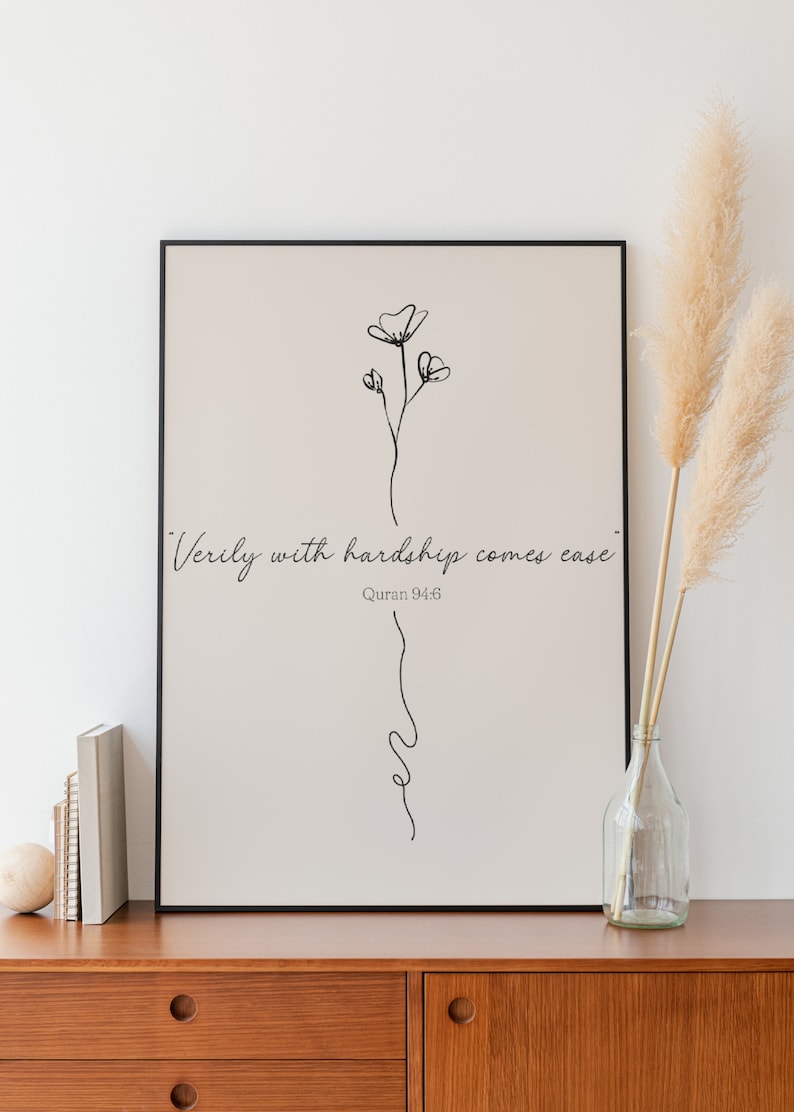 May include: A black framed print with a beige background featuring a simple line drawing of a flower and the quote "Verily with hardship comes ease."  Quran 94:6