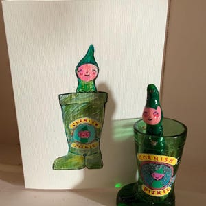 May include: A green glass boot-shaped shot glass and a greeting card featuring a drawing of a Cornish Piskie. The card and glass both display the words "Cornish Piskie". The Piskie is a small, green-clad figure with a pink face.