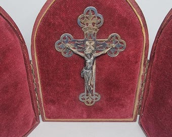 May include: A silver crucifix with a detailed figure of Jesus Christ is displayed inside a red velvet case. The cross has ornate detailing and is set against a deep red background. The case is open, revealing the cross.