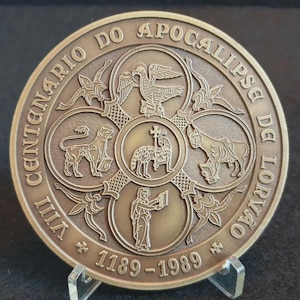 Puede incluir: Una moneda conmemorativa de color bronce con el texto "VIII CENTENARIO DO APOCALIPSE DE LORVÃO 1189-1989". La moneda presenta imágenes religiosas, incluyendo un cordero, un toro, un águila y un león, dentro de un diseño circular.