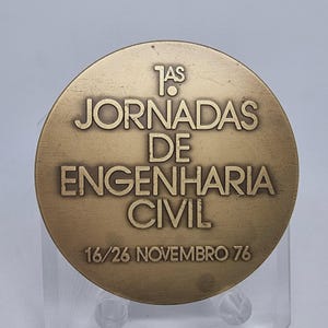 May include: A round, bronze-colored medal with the text "1as JORNADAS DE ENGENHARIA CIVIL" and "16/26 NOVEMBRO 76" engraved on it. The medal has a slightly worn appearance.