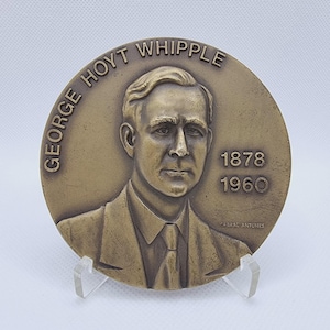 Puede incluir: Medalla conmemorativa de color bronce que presenta a George Hoyt Whipple, con los años 1878 y 1960. El texto "GEORGE HOYT WHIPPLE" está inscrito en el borde superior. El nombre del artista, CABRAL ANTUNES, está en la parte inferior.