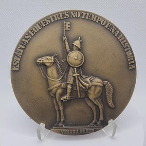 Peut inclure: Médaillon rond de couleur bronze représentant un chevalier à cheval, tenant une lance en forme de clé et un bouclier. Le texte "ESTÁTUAS EQUESTRES NO TEMPO E NA HISTÓRIA" est inscrit sur le bord. Le médaillon est présenté sur un support transparent.